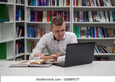 Handsome blond guy in the library working on a laptop in front of him and opened book on the desk with bookshelf full of books in the background