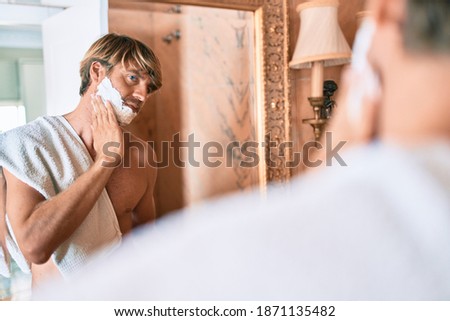 Handsome blond man looking at the mirror at the bathroom saving using foam and razor
