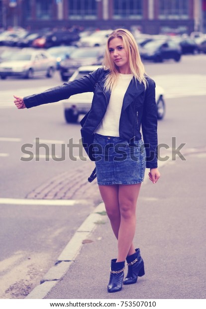 A\
handsome blonde girl needs a lift to another district of a city. So\
she is trying to pick up a car. She is wearing denim skirt and\
black leather coat. With a tiny smile on her\
face.