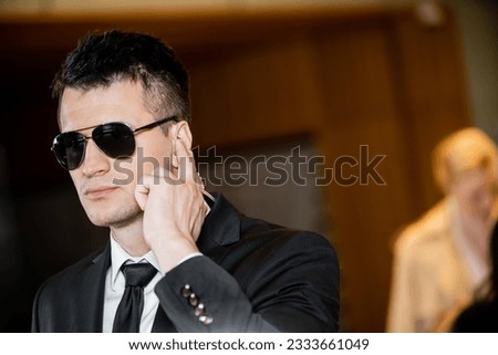 handsome bodyguard in dark sunglasses, handsome man in suit and tie touching earpiece in lobby of hotel, security and career, communication, vigilance, private safety, hotel safety, male personnel
