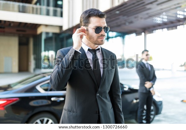 Handsome bodyguard listening to earpiece with car\
parked on street in\
city