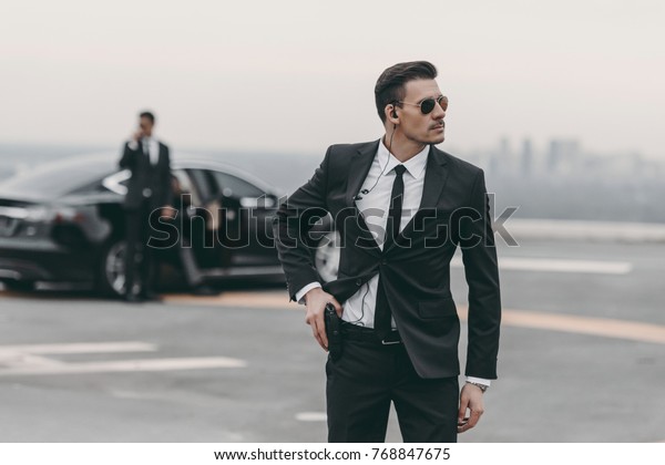 handsome bodyguard with security earpiece putting\
hand on gun