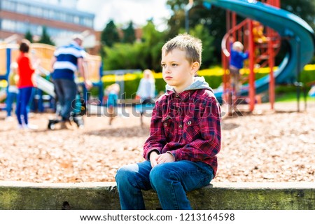 A handsome boy with ADHD, Autism, Asperger Syndrome sitting at the park, scared and apprehensive about playing with the other children, looking nervous and worried