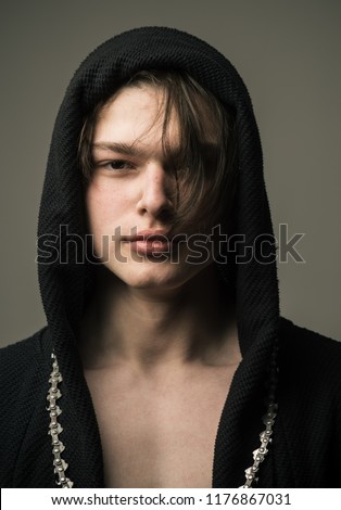Handsome boy in black hood with medium length layered dark hair with long chain around his neck standing isolated on gray background, youth fashion concept.