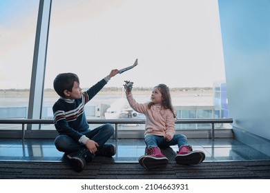 Handsome Boy And Girl, Brother And Sister Playing With Toy Airplanes Sitting On The Floor By The Panoramic Windows Of The Airport Departure Terminal Overlooking The Runways. Family Air Travel Concept