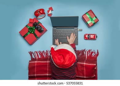 Handsome Boy In Santa Hat On The Blue Floor With A Laptop Xmas Gifts And Toy Car. Merry Christmas And New Year Present. Happy Holidays At Home. Online Shopping By Laptop. Media Addiction. Modern Youth