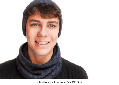 A Handsome Boy In A Warm Winter Hat And A Bondan Scarf On A White Background With Braces On His Teeth. He Smiles. Isolated