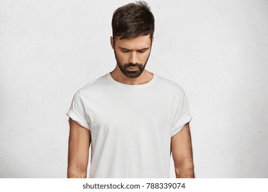 Handsome brunet male with trendy hairdo, has muscular body, wears caual white t shirt, looks down as notices something on floor, poses against concrete background. Attractive man hangs head.