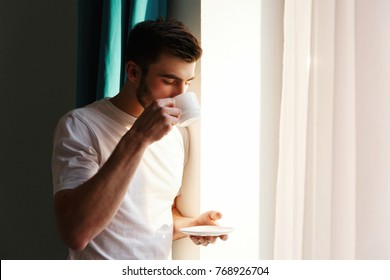 Handsome brunette bearded man stands before bright window and drinks coffee - Powered by Shutterstock