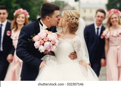 Handsome brunette groom kissing beautiful bride in wedding dress with bouquet bridesmaids in background