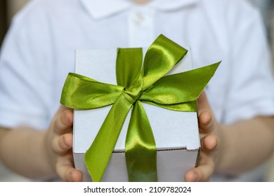 Handsome Brunette Hair Boy Is Holding A White Gift Box With Green Color Ribbon In His Hands. Cute Kid In White T Shirt Is Smiling Or Has Serious Face. Free Space, For Greeting Card, Womans Or Mother's