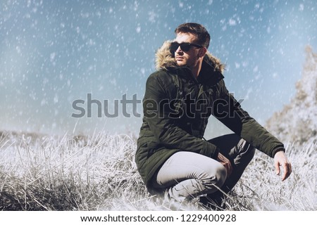 A handsome brutal man wearing a parka and sunglasses in the countryside. Fashion for men. Autumn, winter.
