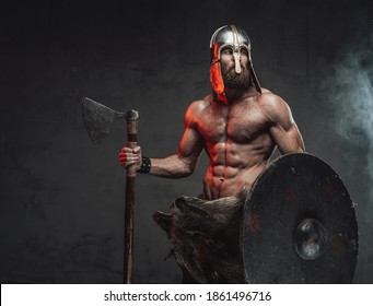 Handsome and brutal viking with beard and naked torso with helmet on him posing staying with axe and shield in smoky dark background.