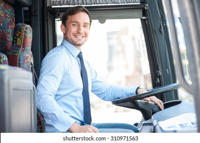 Handsome bus driver is sitting at steering wheel. He is looking at the camera and smiling