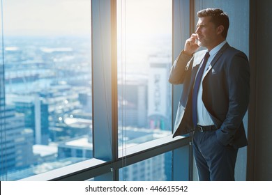 Handsome business CEO with designer stubble, talking confidently on his mobile phone while looking out of large windows in a top floor office at the city below