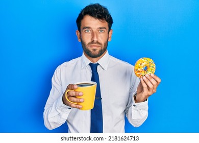 Handsome business man with beard eating doughnut and drinking coffee relaxed with serious expression on face. simple and natural looking at the camera. 