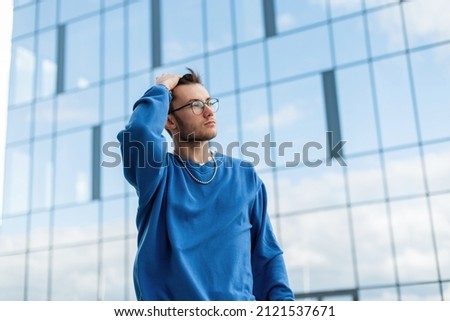 Handsome business man model hipster with hairstyle in fashion blue sweatshirt walks in the city near a modern glasses building