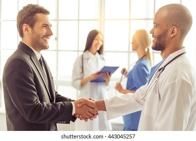 Handsome businessman and Afro American doctor are shaking their hands and smiling, two female doctors are talking in the background - Powered by Shutterstock
