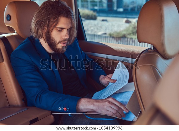 A\
handsome businessman with a beard and long hair sitting in the back\
seat of a luxury car and working with\
documents.