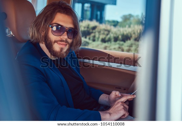 A handsome businessman with a beard\
and long hair sitting in the back seat of a luxury\
car.