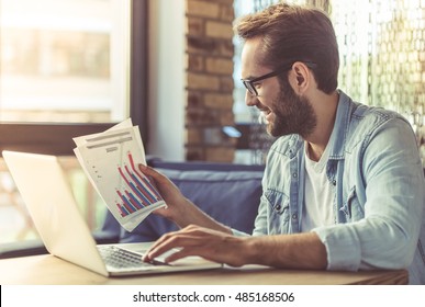 Homem de negócios bonito em roupas casuais e óculos está usando um laptop, examinando documentos e sorrindo enquanto trabalha no escritório