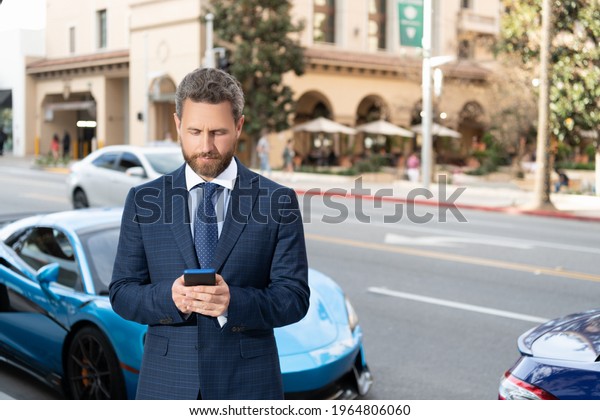 handsome businessman chatting on smartphone stand\
by luxury auto outdoor. car available. man in expensive suit near\
automobile. car insurance business. successful dealer. confident\
manager rent online