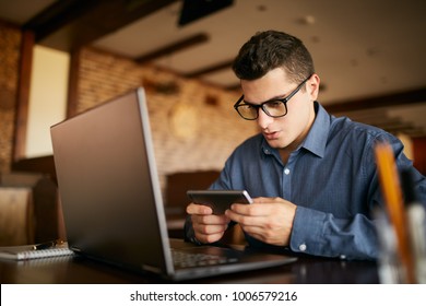 Handsome Businessman Distracted From Work On The Laptop Watching Video On Smartphone. Freelancer Holding Mobile Phone And Browsing Using High Speed 4g Or 5g Internet. Man Playing Mobile Games At Work