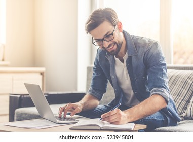 Handsome businessman in eyeglasses is making notes and smiling while working with a laptop at home - Powered by Shutterstock