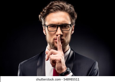 Handsome businessman in eyeglasses and suit gesturing for silence with finger isolated on black - Shutterstock ID 548985511