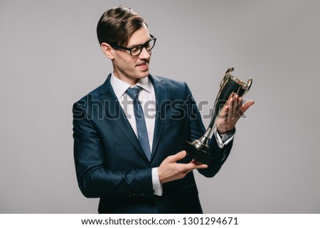 handsome businessman in glasses looking at trophy isolated on grey