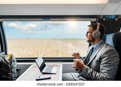 Handsome businessman is having a good time while traveling by high-speed-train. He is using laptop computer and wireless headphones for online communication, gaming and entertainment.