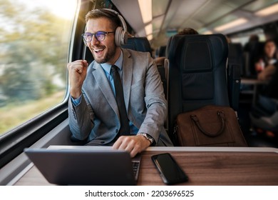 Handsome businessman is having a good time while traveling by high-speed train. He is using laptop computer and wireless headphones for online communication, gaming and entertainment.