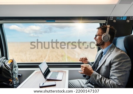 Handsome businessman is having a good time while traveling by high-speed-train. He is using laptop computer and wireless headphones for online communication, gaming and entertainment.