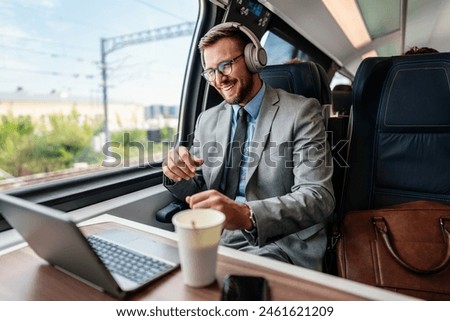 Handsome businessman is having a good time while traveling by high-speed train. He is using laptop computer and wireless headphones for online communication, gaming and entertainment.