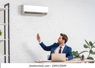 handsome businessman holding remote control while sitting at workplace under air conditioner - Powered by Shutterstock