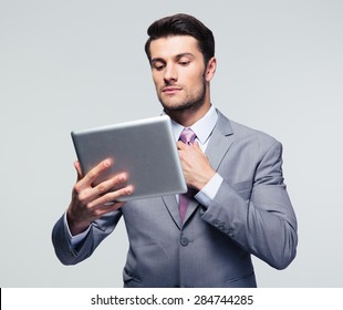 Handsome businessman holding tablet computer and straightening his tie over gray background