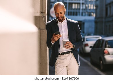 Handsome Businessman Leaning On A Wall While Standing Outdoors And Using Smartphone. Man In Suit Wearing Earphones Looking At His Mobile Phone.