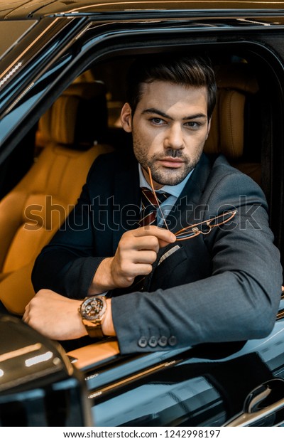 handsome businessman with luxury watch holding\
eyeglasses while sitting in\
car