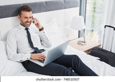 Handsome Businessman Making Phonecall While Sitting On Hotel Room Bed And Using Laptop During Business Trip