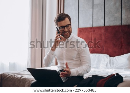 Handsome businessman making phonecall while sitting on hotel room bed and using laptop during business trip