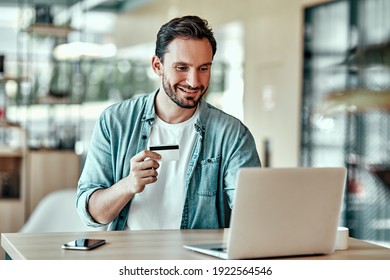 Handsome businessman shopping online with credit card. Portrait of smiling man using laptop computer in cafe - Shutterstock ID 1922564546