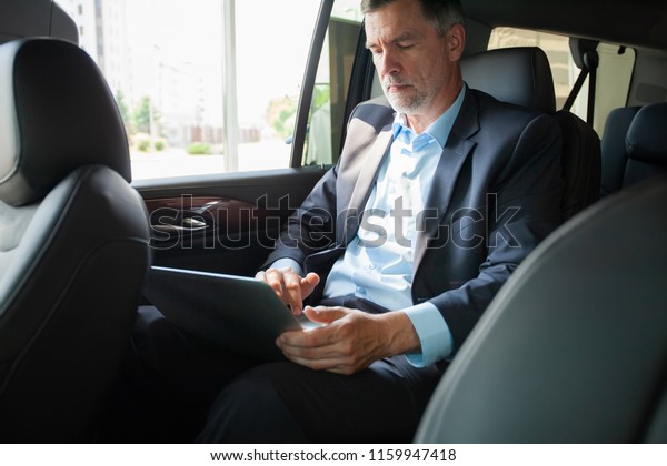 Handsome businessman sitting with\
laptop on the backseat of the car. Caucasian male business\
executive travelling by a car and working on laptop\
computer