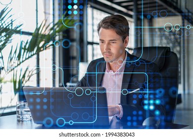 Handsome Businessman In Suit At Workplace Working With Laptop To Optimize Development By Implying New Technologies In Business Process. Hi Tech Hologram Over Office Background