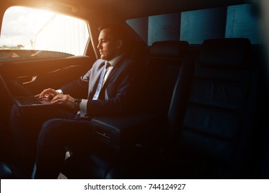 Handsome businessman talking with phone sitting with laptop on the backseat of the car