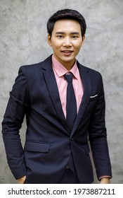 Handsome Businessman Wearing Pink Shirt, Tie, Suit In Black, Standing Smiling, Looking Camera, Right Hand In Pant Pocket. Photo Id Shooting In Studio With A Loft Background