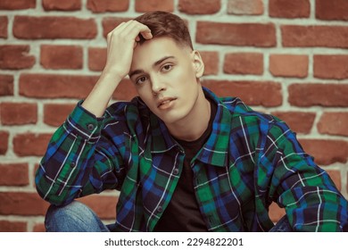 A handsome calm young man in a flannel plaid shirt against a brick wall. Youth fashion. People and emotions. Studio portrait.