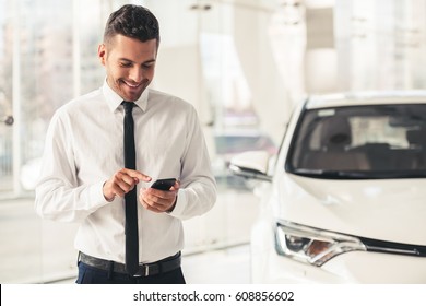 Handsome Car Dealership Worker In Suit Is Using A Smart Phone And Smiling While Standing Near The Car