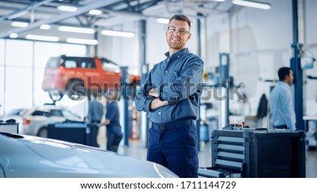 Handsome Car Mechanic is Posing in a Car Service. He Wears a Jeans Shirt and Safety Glasses. His Arms are Crossed. Specialist Looks at a Camera and Smiles.