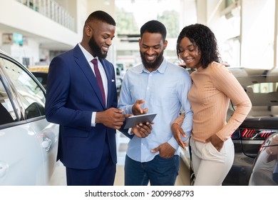 Handsome Car Seller Showing Purchasing Details On Digital Tablet To Young Black Couple In Dealership Center, Happy African American Spouses Buying New Vehicle In Automobile Showroom, Closeup