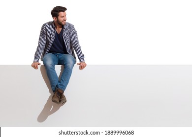 Handsome casual young man is sitting on a top of white banner with legs crossed, smiling and looking at the side. Full length studio shot isolated on white.
